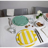 Four round striped table placemats in green, blue, yellow, and red are arranged on a white tablecloth with silver cutlery, glasses, and a small white flower centerpiece.