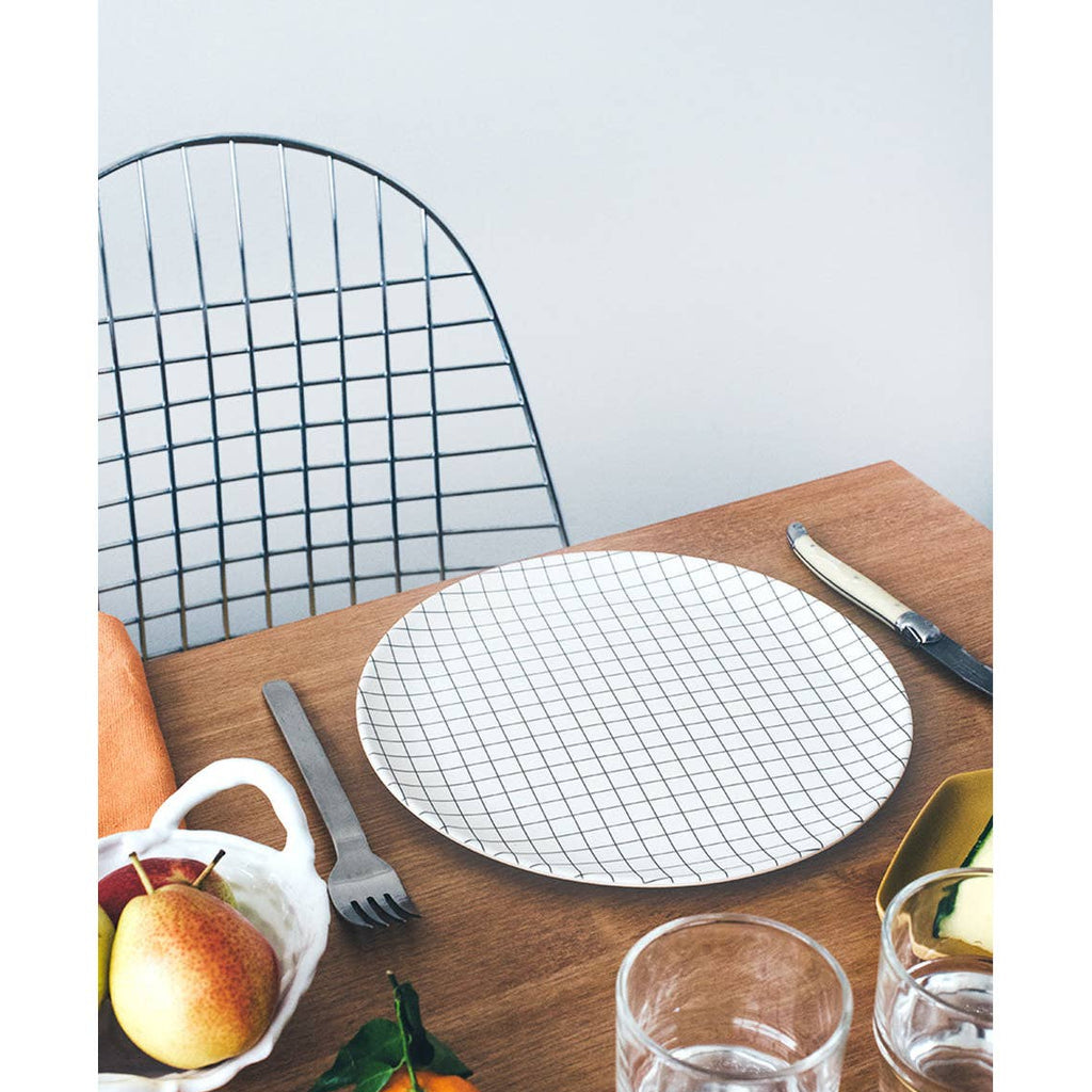 A round white placemat with a black grid pattern is set on a wooden dining table, surrounded by a metal chair, silver cutlery, glasses, and small fruit and vegetable accents.