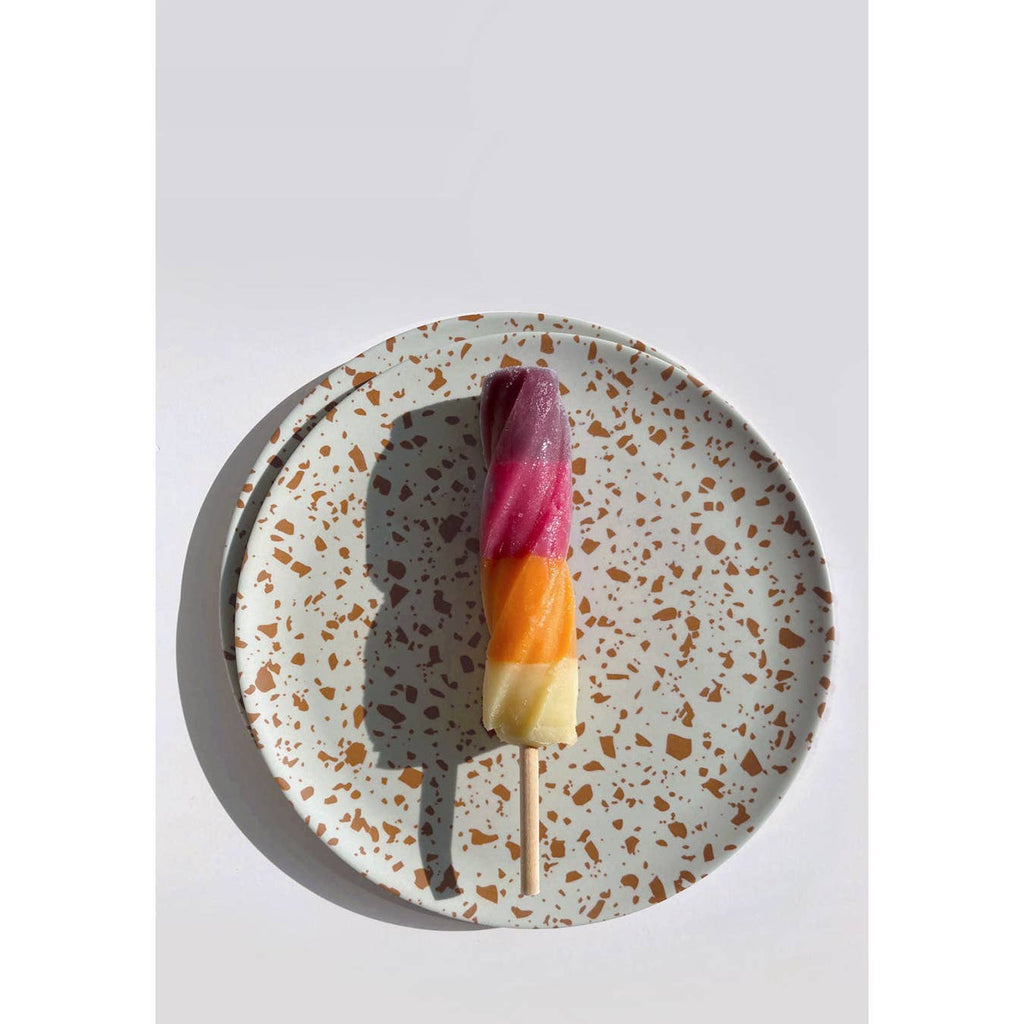 A three-colored popsicle with purple, orange, and yellow layers on a wooden stick, presented on a speckled white ceramic plate against a light gray background.