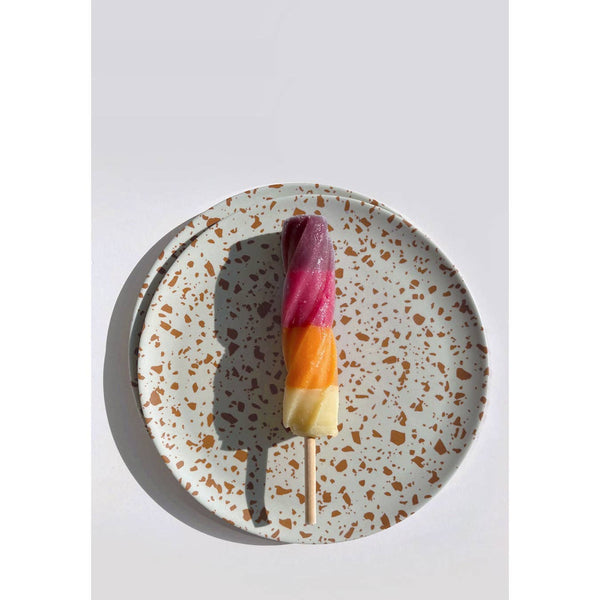 A three-colored popsicle with purple, orange, and yellow layers on a wooden stick, presented on a speckled white ceramic plate against a light gray background.