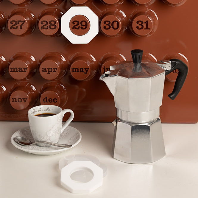 Stovetop espresso maker with a cup of coffee on a white surface against a brown wall with numbered tiles.