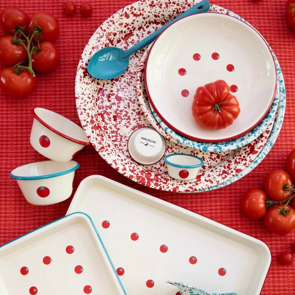A styled flat-lay of a red, white, and blue speckled enamel dinnerware set on a red checkered tablecloth, surrounded by fresh tomatoes. The set includes a large rectangular tray, a round tray, a small cup, a bowl, a plate, a spoon, and a small pitcher.