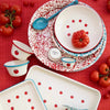 A styled flat-lay of a red, white, and blue speckled enamel dinnerware set on a red checkered tablecloth, surrounded by fresh tomatoes. The set includes a large rectangular tray, a round tray, a small cup, a bowl, a plate, a spoon, and a small pitcher.