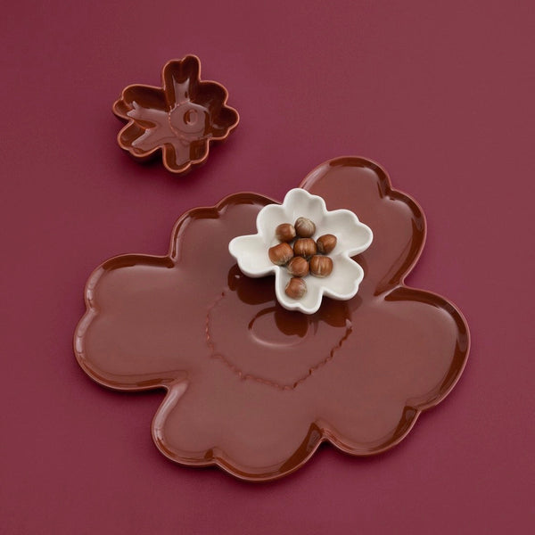 A flower-shaped brown ceramic serving platter with a matching small flower-shaped dish, shown on a dark burgundy background. A small white flower dish in the center holds brown nuts.