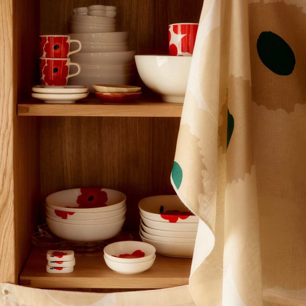 A kitchen cabinet with wooden shelves holding white ceramic dishes with red poppy motifs, alongside a beige curtain with large green and cream abstract shapes.