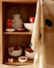 A kitchen cabinet with wooden shelves holding white ceramic dishes with red poppy motifs, alongside a beige curtain with large green and cream abstract shapes.