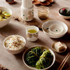 A styled Japanese dining table setting featuring a ceramic tableware set with a floral pattern, including bowls, a cup, a small pitcher, and a teapot, arranged with food on a rustic wooden table.
