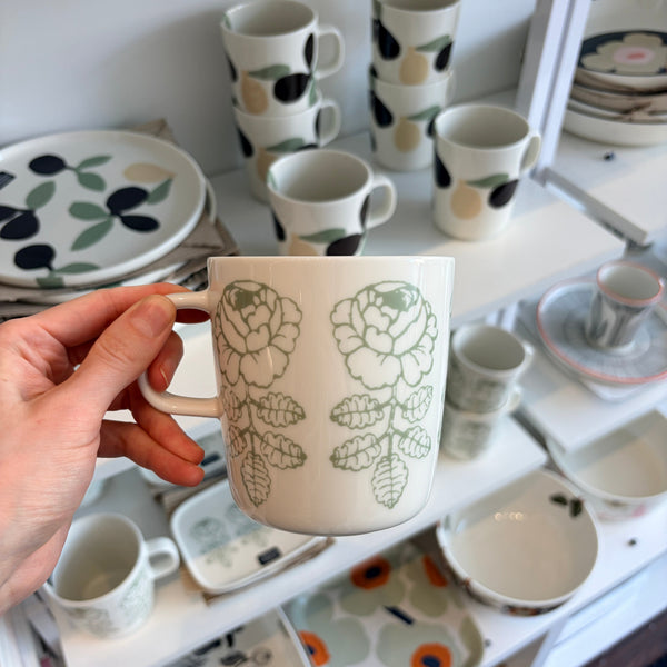 Hand holding a floral mug with a background of variously decorated mugs and plates.