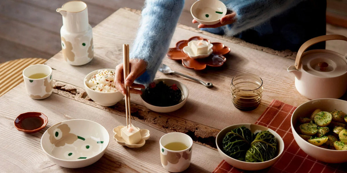 A person in a cream and blue gradient sweater is shown from the waist up, using chopsticks to serve food from a rustic wooden table set with various ceramic dishes, bowls, and cups.
