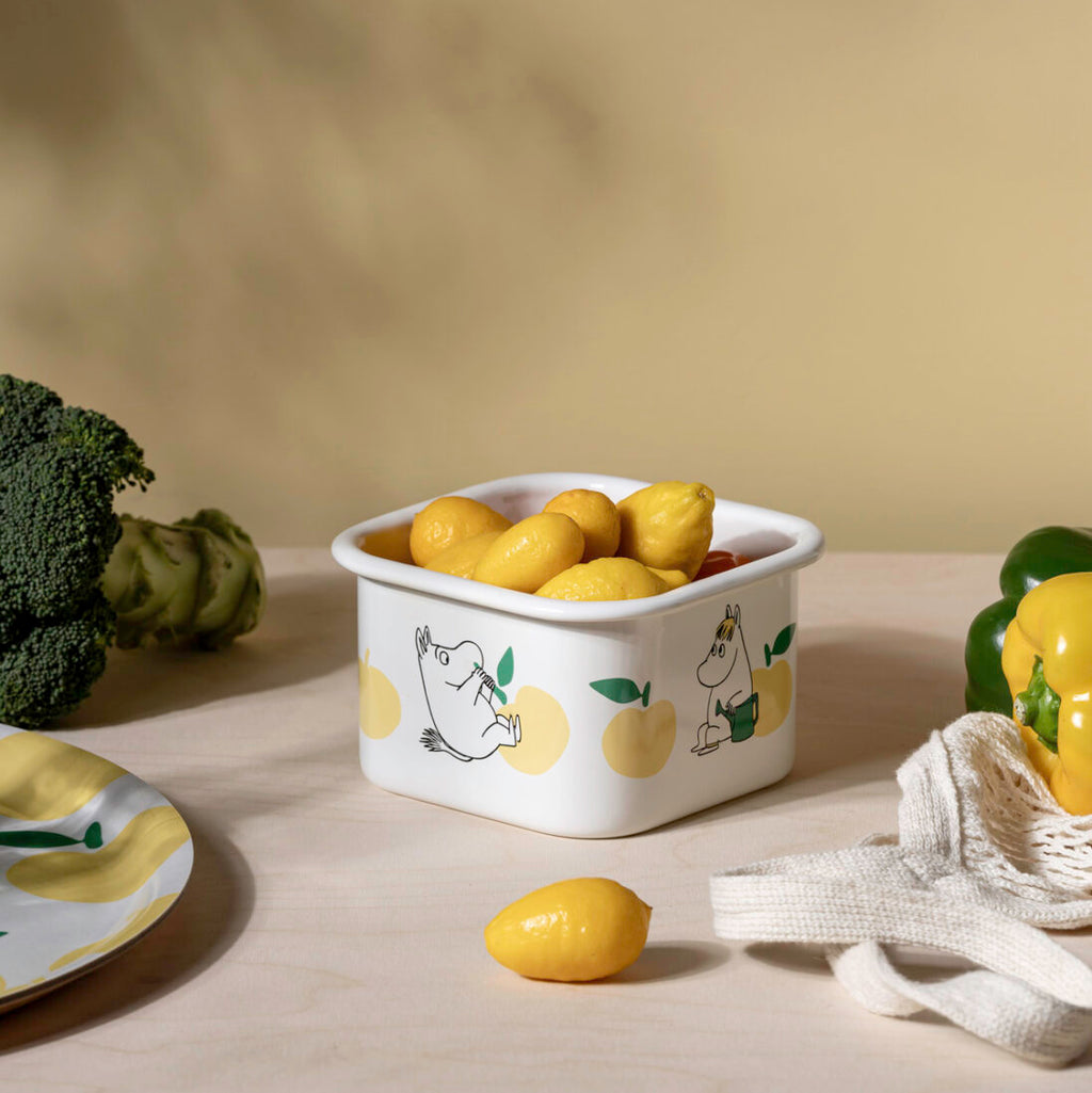 White container with lemon design on a wooden surface with lemons and a towel.