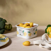 White container with lemon design on a wooden surface with lemons and a towel.