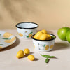 Two small ceramic bowls with black rims, one containing lemons and the other with green apples, on a beige surface.