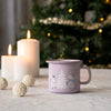Purple mug with illustrations on a table with candles and a decorated Christmas tree in the background.