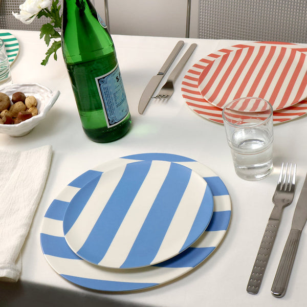 A styled dining table setting featuring blue and white striped ceramic plates, a green glass bottle, silver cutlery, a clear drinking glass, a folded white napkin, and a small white flower on a light neutral background.